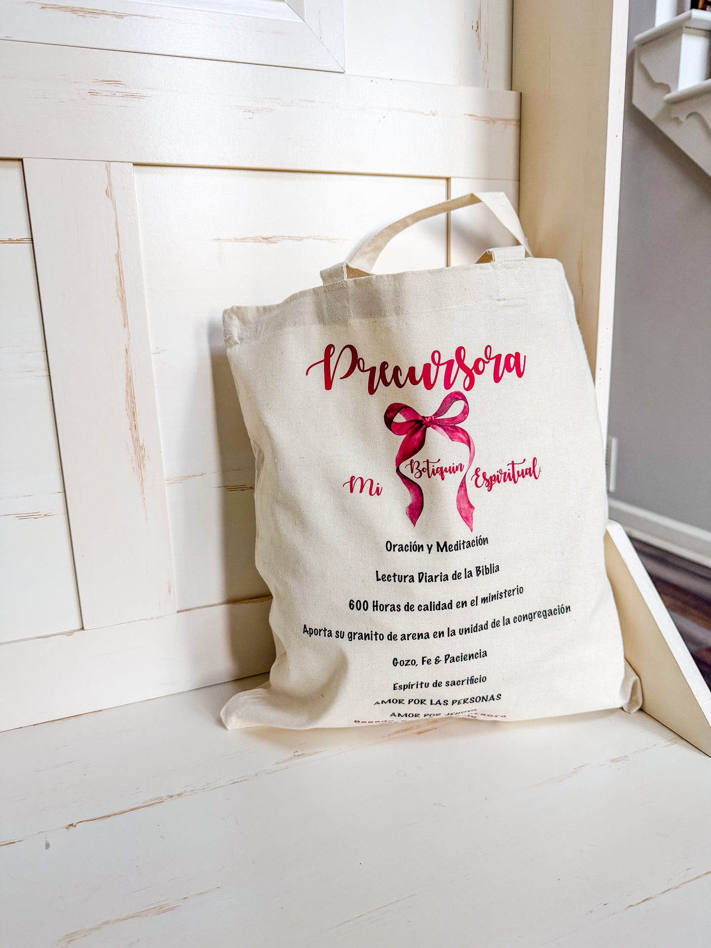 Beige tote bag with red text and a pink bow on a white surface.